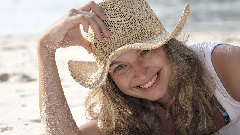 Blonde, lachende Frau am Strand mit Strohhut