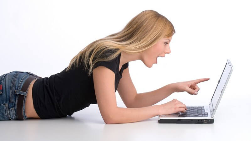 Blondes Mädchen liegt auf dem Bauch vor einem Laptop, zeigt auf den Bildschirm und hat den Mund geöffnet