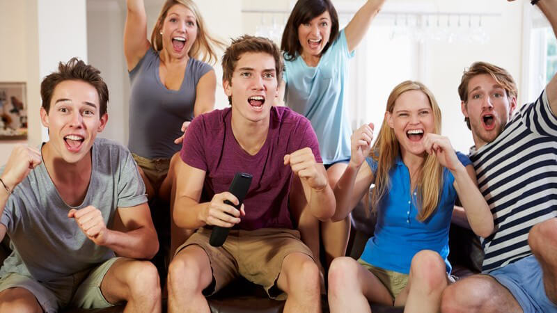 Sechs jugendliche Freunde sitzen mit Popcorn vor dem Fernseher und jubeln