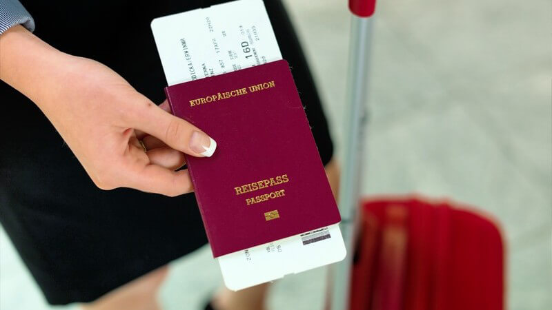 Frau mit rotem Trolley hält einen Reisepass mit inliegendem Flugticket hin