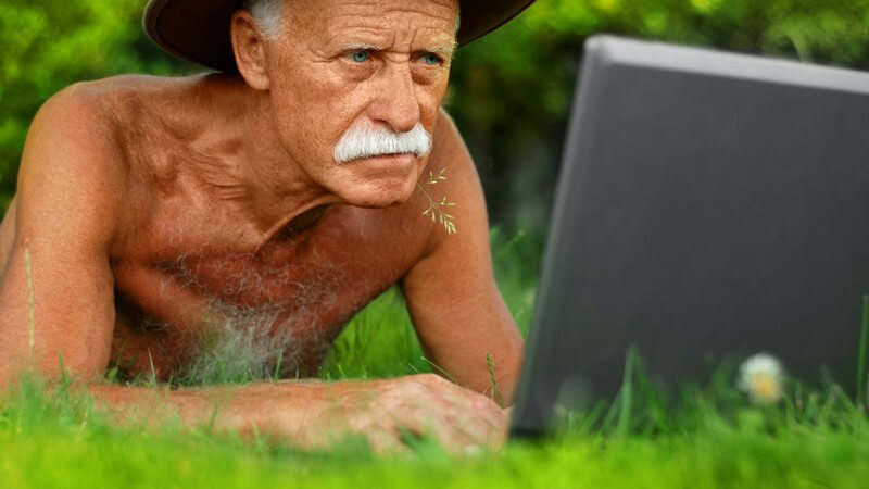 Alter Mann mit weißem Bart und Hut liegt oberkörperfrei auf Wiese mit aufgeklapptem Laptop