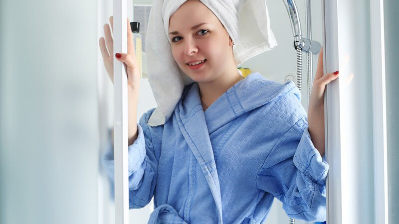 Frau in Bademantel mit Handtuch auf Kopf steht in Dusche