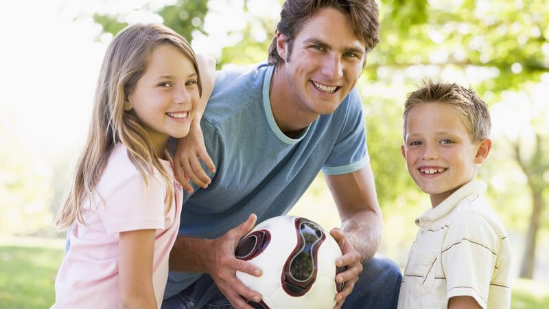 Vater mit Tochter und Sohn draußen mit Volleyball, alle lachen in Kamera