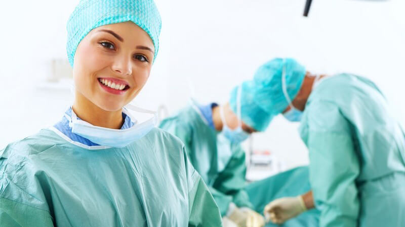 Ärztin lächelt in die Kamera, im Hintergrund Ärzte bei der Operation