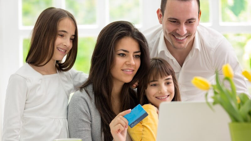 Junge Familie sitzt gemeinsam vor einem Laptop, die Mutter hält eine blaue Kreditkarte in der Hand