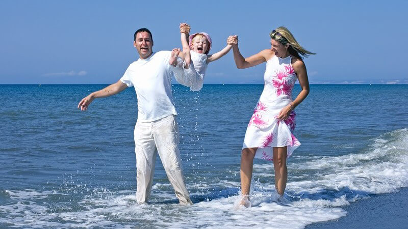 Familie am Strand, Vater in weiß, Mutter in weiß-pinkem Kleid, mit Tochter o. Mädchen in der Mitte in Wasser bei Sonne