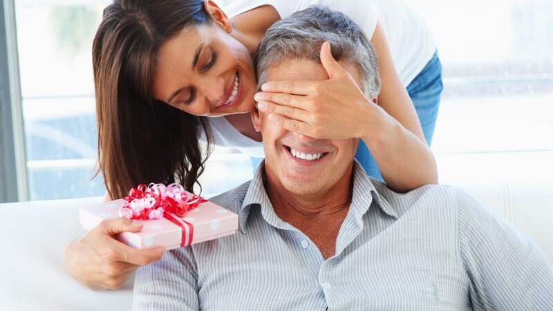 Frau hält Mann von hinten Augen zu, hält Geschenk in Hand, Überraschung
