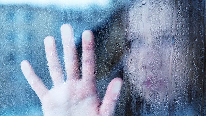 Scheibe mit Regentropfen, im Hintergrund Gesicht einer traurigen Frau mit Handfläche auf Scheibe