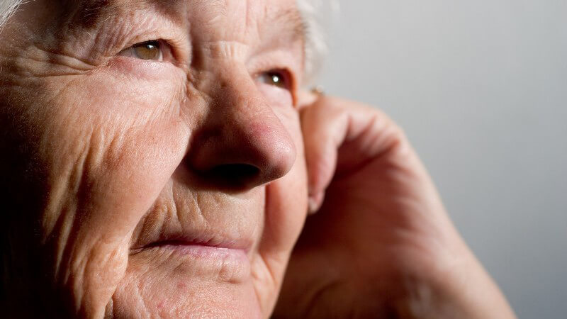 Alte Frau mit braunen Augen blickt in die Ferne, die linke Hand an der Wange