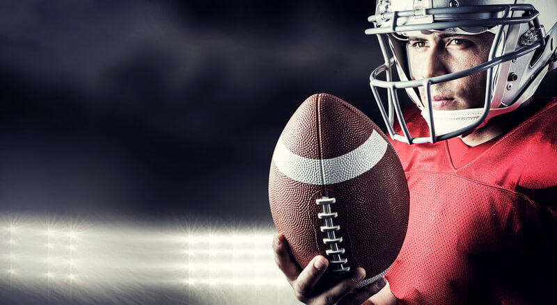 American Football-Spieler in rotem Trikot und weißem Helm hält einen Football in der Hand, Flutlicht im Hintergrund