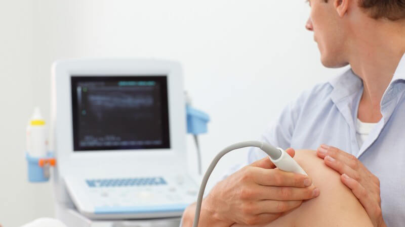 Junger Arzt untersucht ein Knie per Ultraschallgerät und schaut auf den Monitor