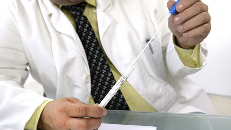 Laborant in weißem Kittel und Krawatte sitzt am Tisch und hält ein Abstrichröhrchen zur DNA-Analyse in den Händen