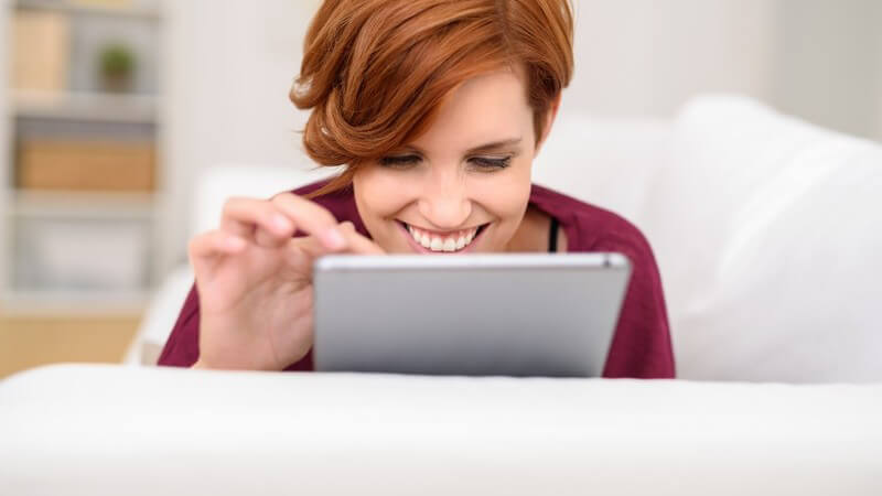 Junge Frau mit kurzen Haaren liegt auf einer weißen Couch und tippt lächelnd auf ihrem Tablet-PC