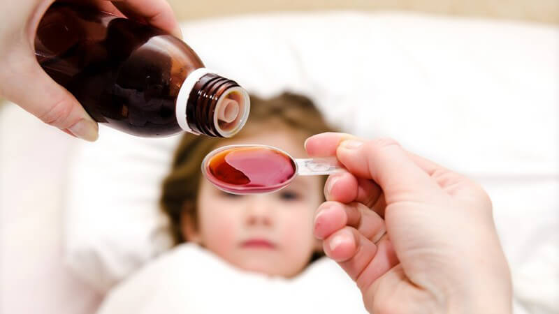 Flüssige Medizin wird auf einen Löffel gegeben, im Hintergrund ein krankes Kind im Bett