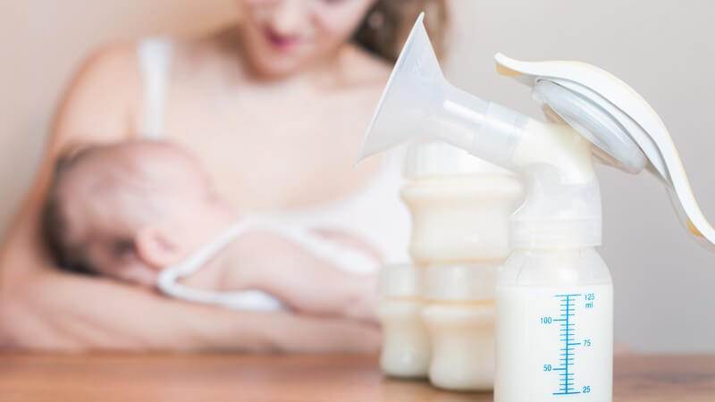 Manuelle Milchpumpe auf Tisch, im Hintergrund sitzt eine Mutter mit ihrem Baby im Arm