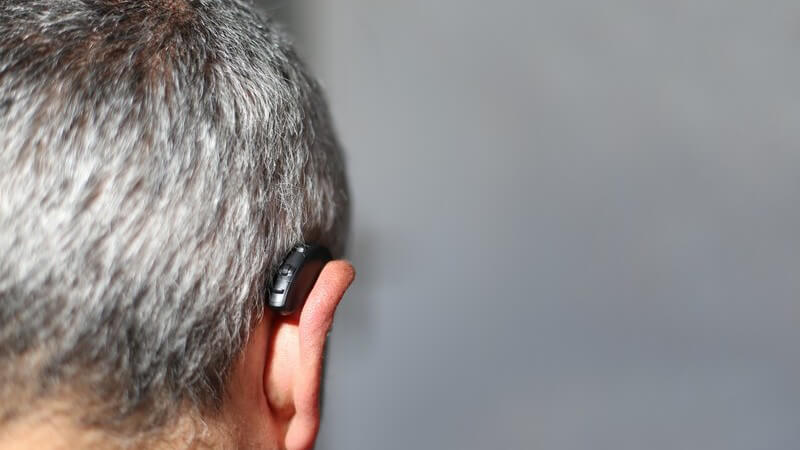 Rückansicht des Kopfes eines grauhaarigen Mannes mit einem Hörgerät im Ohr, grauer Hintergrund