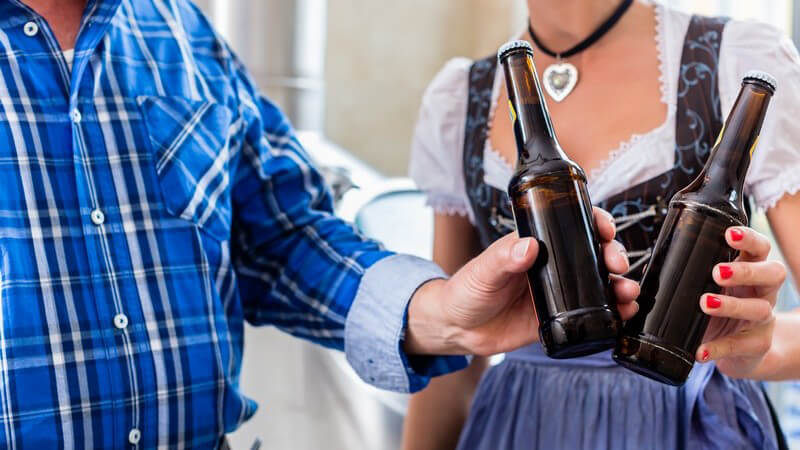 Braumeister in blauem Hemd und Frau im Dirndl stoßen mit Bierflaschen an