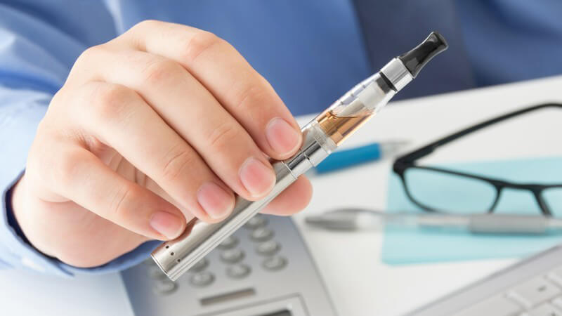 Mann hält eine silberne E-Zigarette über dem Schreibtisch mit Taschenrechner, Brille und Tastatur