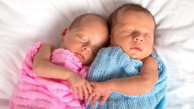 Neugeborene Zwillinge schlafen eng aneinander in rosa und hellblauem Wollsack
