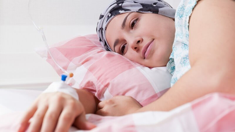 Junge Frau mit grauem Kopftuch (Chemotherapie) liegt zuversichtlich im Krankenhaus, auf dem Handrücken ein Zugang