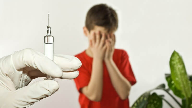 Hand eines Arztes hält Spritze in Kamera, im Hintergrund ängstlicher Junge