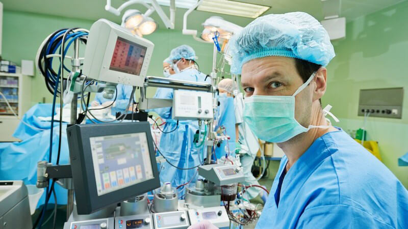 Arzt in blauer Operationskleidung sitzt im OP-Saal an diversen Geräten, im Hintergrund läuft eine Operation