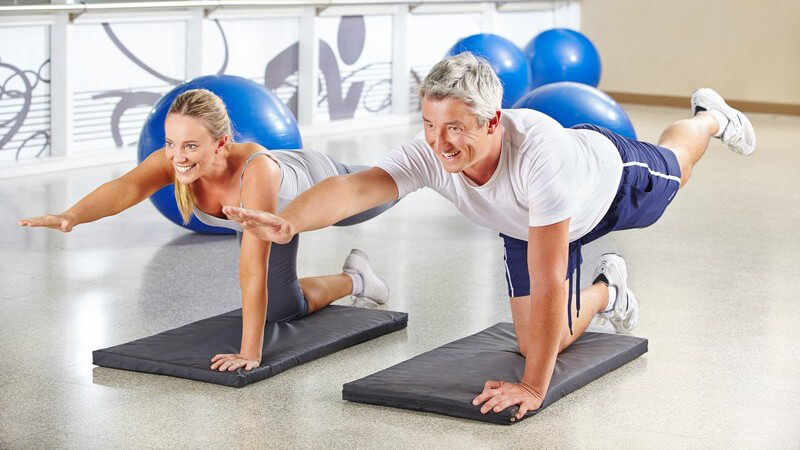 Frau und älterer Mann machen Gymnastik auf grauen Matten, im Hintergrund liegen blaue Gymnastikbälle