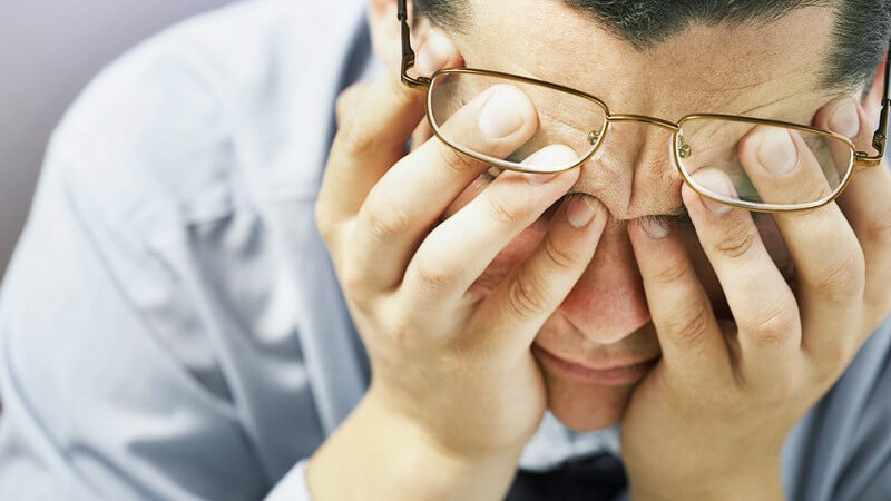 Mann in Hemd und Krawatte reibt sich die Augen unter seiner Brille, wirkt erschöpft oder depressiv