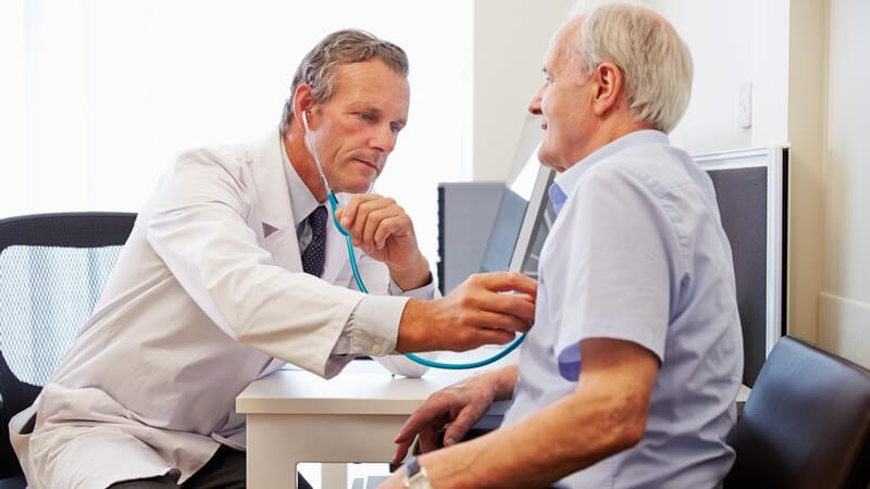 Älterer Mann in blauem Hemd sitzt bei einem Arzt in der Sprechstunde und wird mit dem Stethoskop abgehört