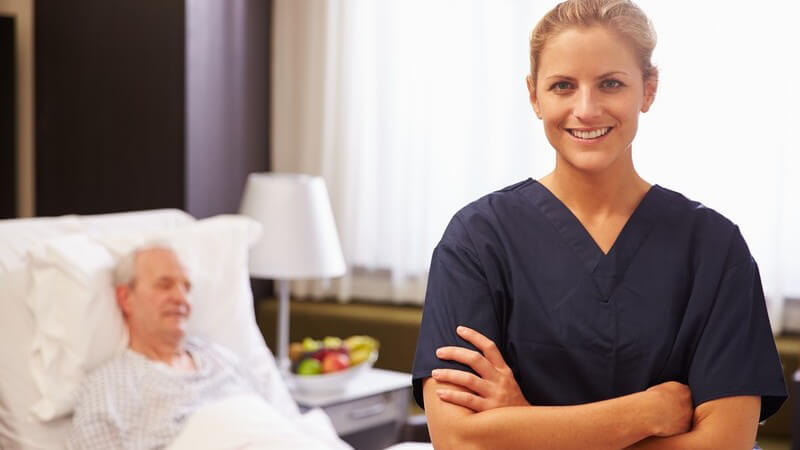 Blonde Krankenschwester posiert in dunkelblauem Kasack vor einem alten Mann im Krankenbett