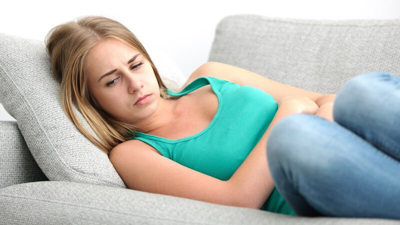 Junge Frau in Jeans und türkisem Oberteil krümmt sich mit Bauchschmerzen auf einem grauen Sofa