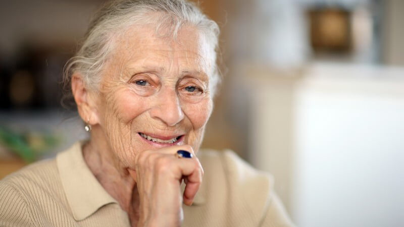 Alte Dame lächelt, das Kinn auf die rechte Hand gestützt, blauen Ring am Ringfinger