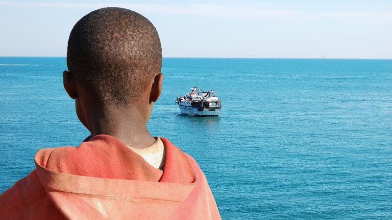 Dunkelhäutiger Flüchtlingsjunge in rotem Kapuzenpullover blickt aufs offene Meer in Richtung eines großen Schiffes