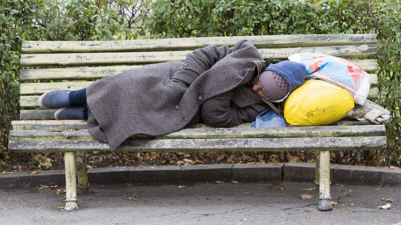Obdachloser schläft auf einer Parkbank, gelbe Tüte als Kissen