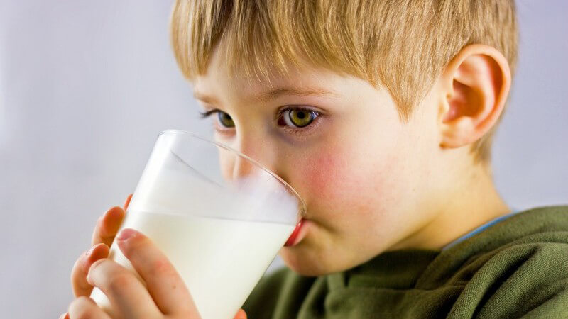 Kleiner, dunkelblonder Junge in grünem Kapuzenpullover trinkt aus einem Glas mit Milch