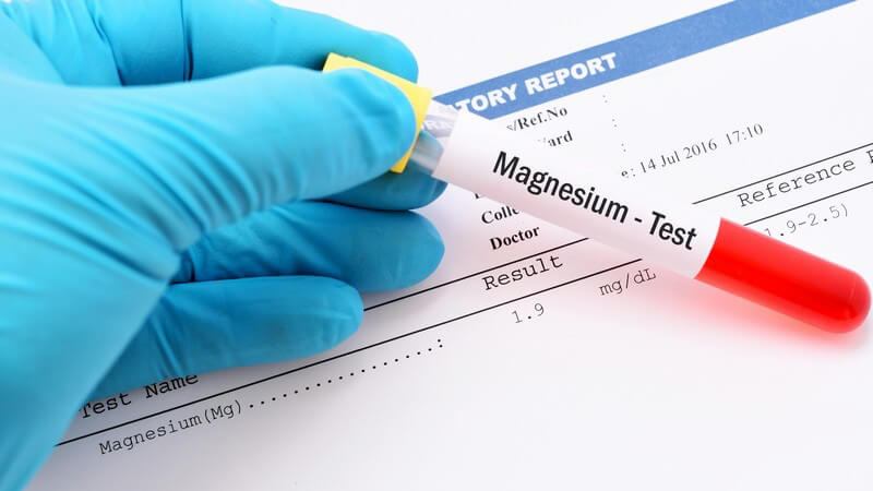 Hand mit blauem Handschuh hält ein Röhrchen mit einer Blutprobe für einen Magnesium-Test über ein Testergebnis