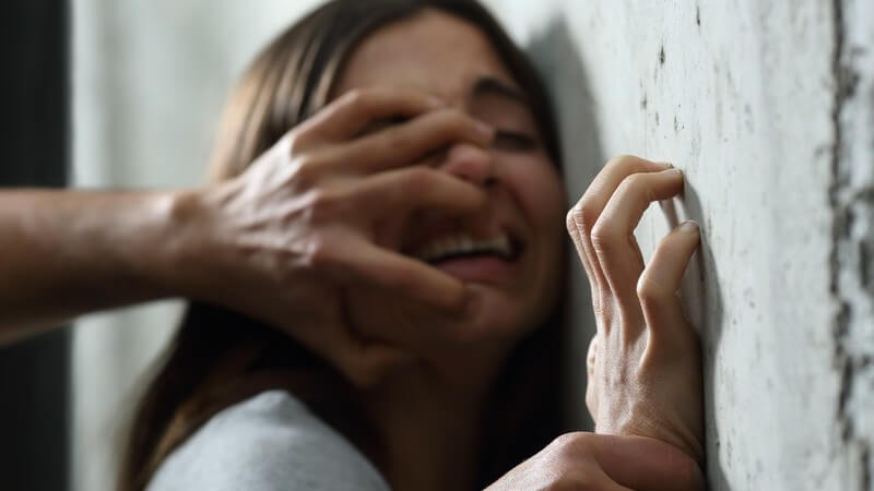 Szene eines sexuellen Missbrauchs, junge Frau wird von hinten attackiert und an Wand gedrückt, die Hand im Gesicht
