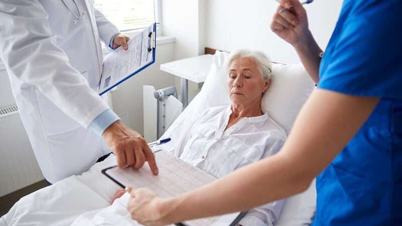 Arztvisite bei einer älteren Patientin im Krankenhausbett, Arzt und Krankenschwester halten ein Klemmbrett in der Hand