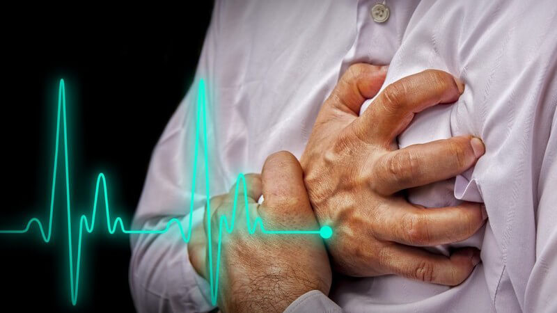 Mann in lila Hemd fasst sich wegen Brustschmerzen an die linke Brust, davor die Herzfrequenz (Fotomontage)