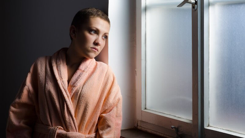 Junge Krebspatientin steht im Bademantel in einem dunklen Raum am Fenster und guckt traurig