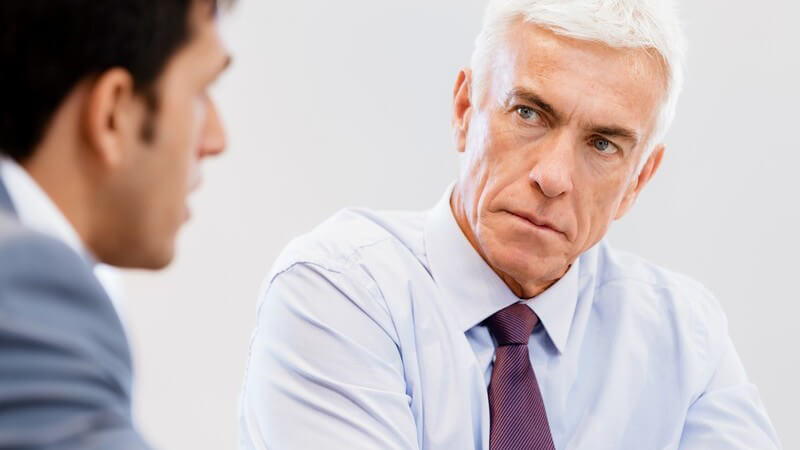 Älterer Geschäftsmann mit weißen Haaren und lila Krawatte guckt ernst und strend während Gespräch mit einem Mitarbeiter