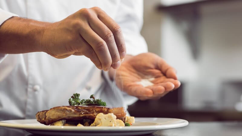 Koch streut mit der rechten Hand Salz über das servierfertige Essen