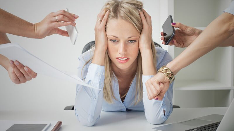 Blonde Geschäftsfrau hängt völlig gestresst auf ihrem Schreibtisch, umgeben von Kollegen-Händen mit Smartphone und Uhr