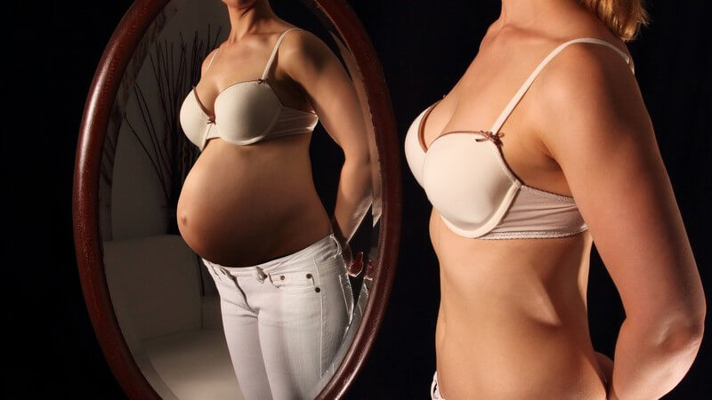 Frau in Hose und BH sieht sich schwanger im Spiegel (Kinderwunsch)