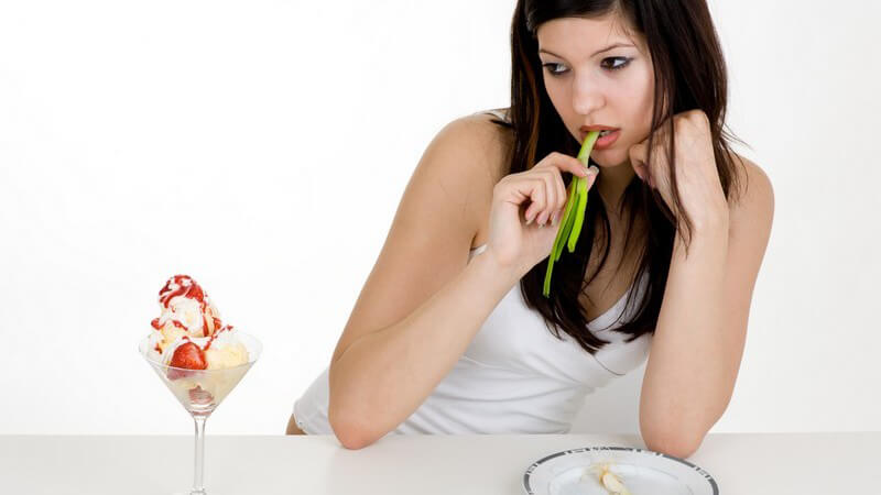 Frau sitzt an Tisch mit Teller und isst Gemüse, schaut neidisch auf Eisbecher neben sich
