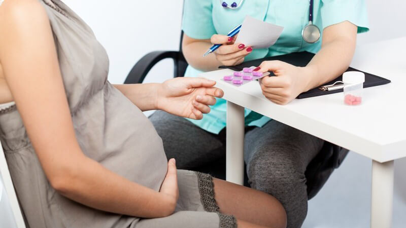 Schwangere Frau in grauem Sommerkleid sitzt beim Frauenarzt und erhält einen Blister mit Tabletten