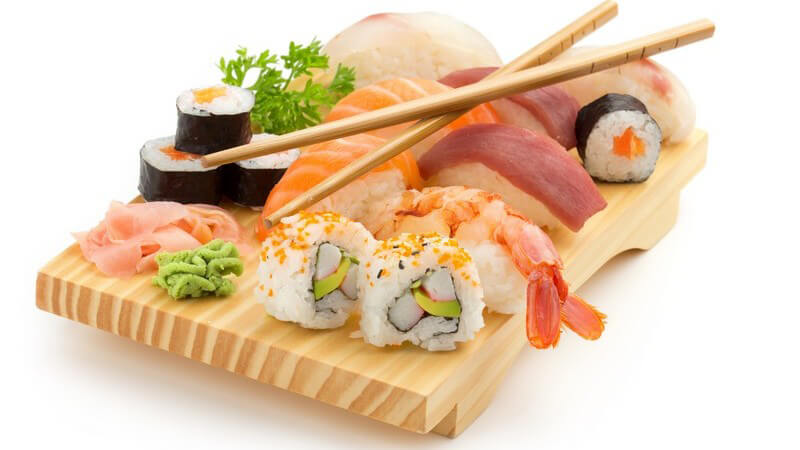 Holzplatte mit diversen Sushi-Arten und Essstäbchen vor weißem Hintergrund