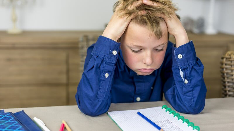 Junge in blauem Hemd sitzt verzweifelt an den Hausaufgaben, die Hände im Haar
