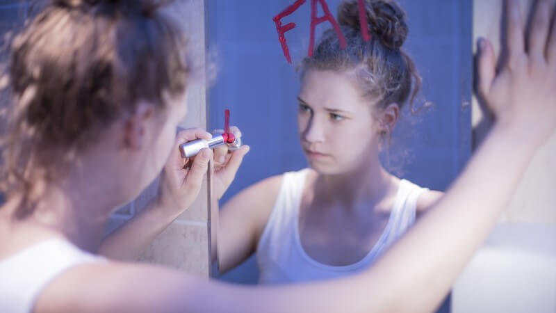 Mädchen schreibt mit einem Lippenstift "FAT" auf einen Spiegel