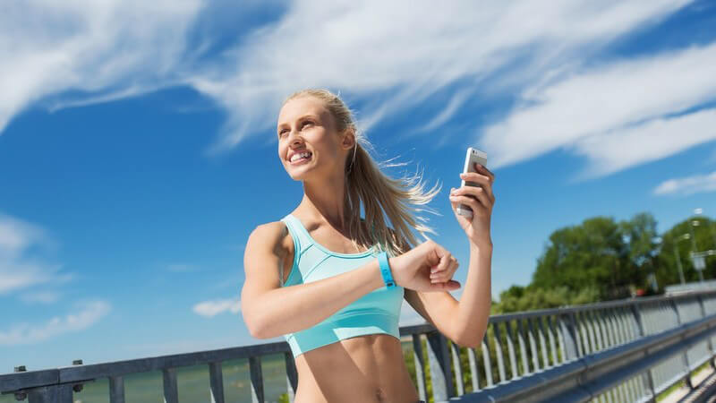 Blonde Joggerin steht auf einer Brücke und checkt ihren Puls mit Fitnessarmband und Smartphone
